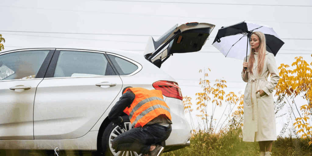 A woman repairs a car while holding an umbrella to shield herself from the rain.